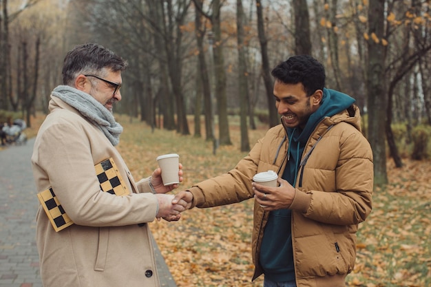 Friends a senior and a young man walking and talking and drinking coffee together in the autumn parkxA