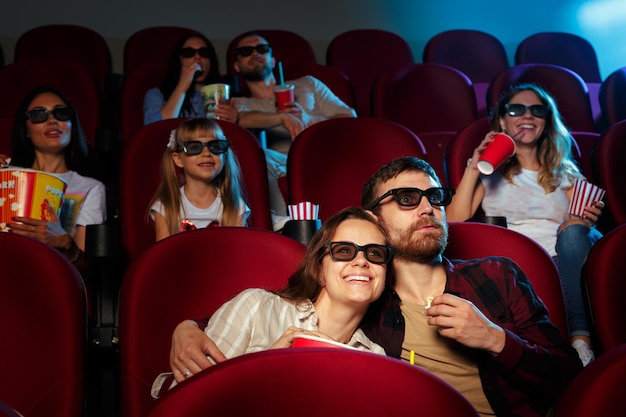 Friends sitting in cinema watch film eating popcorn and drinking water.