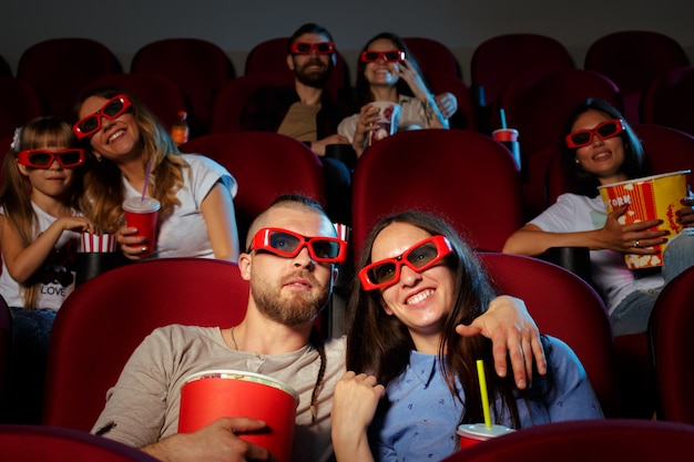 Friends sitting in cinema watch film eating popcorn and drinking water.