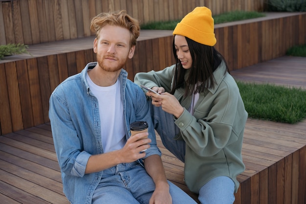 Friends sitting together in park using mobile phone holding cup of coffee