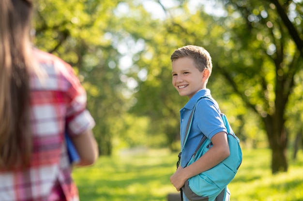 Friends. Smiling boy in blue tshirt with backpack and girl standing with her back to camera outdoors on fine day