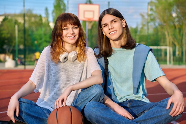 Photo friends teenagers guy and girl looking at camera sitting on basketball court