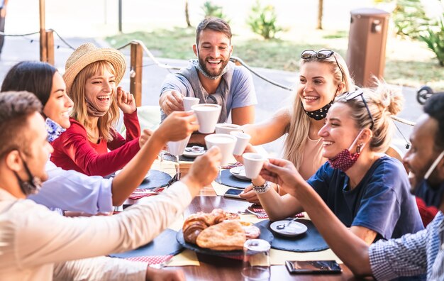 写真 コーヒーショップでカプチーノを乾杯する友人はフェイスマスクを着用します-レストランで一緒に楽しんでいる若者-カフェバーで幸せな男と女との新しい通常のライフスタイルのコンセプト