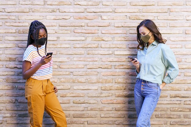 Photo friends wearing mask using smart phone standing by wall outdoors