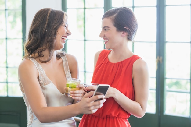 Friends with cocktail glasses laughing while using mobile phone