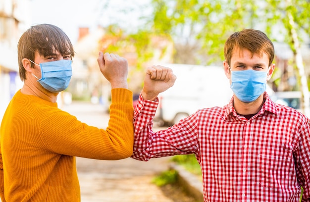 Friends with a protective medical mask on their face greet their elbows