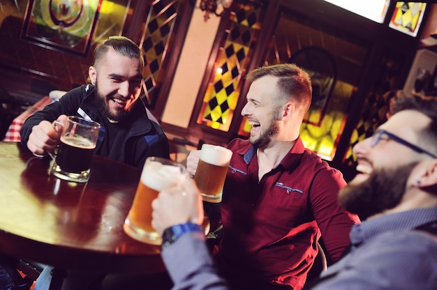 Друзья - молодые симпатичные мужики пьют пиво в баре, звонят в бокалы, улыбаются, смеются и разговаривают.