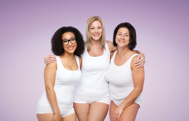 дружба, красота, позитивное тело и концепция людей - группа счастливых женщин больших размеров в белом нижнем белье на фиолетовом фоне