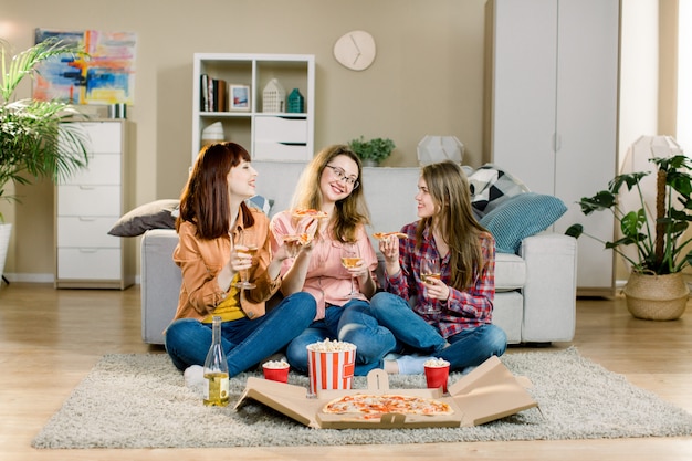 Friendship, holidays, fast food and celebration concept - happy young women friends with drinks and popcorn eating pizza at home, sitting on the floor in cozy room