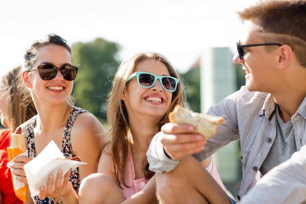 концепция дружбы, отдыха, лета и людей - группа улыбающихся друзей в солнечных очках, сидящих с едой на городской площади