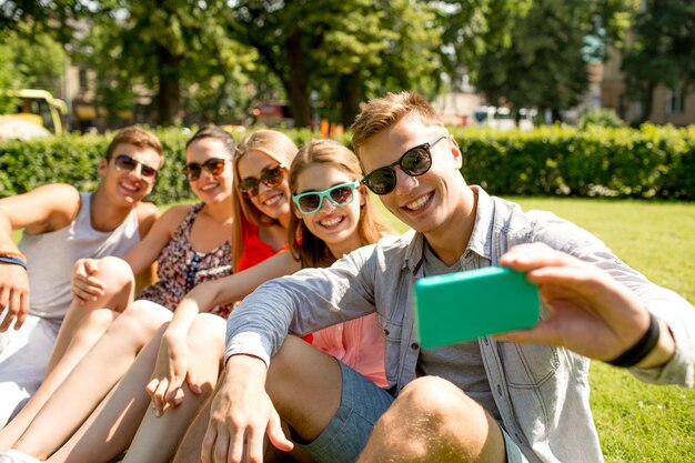 friendship, leisure, summer, technology and people concept - group of smiling friends with smartphone sitting on grass and making selfie in park