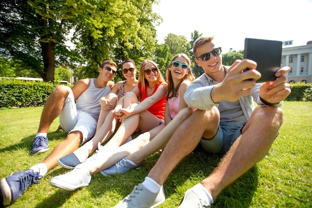 friendship, leisure, summer, technology and people concept - group of smiling friends with tablet pc computer sitting and making selfie in park