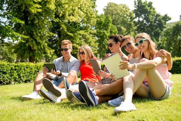 friendship, leisure, summer, technology and people concept - group of smiling friends with tablet pc computers sitting on grass in park