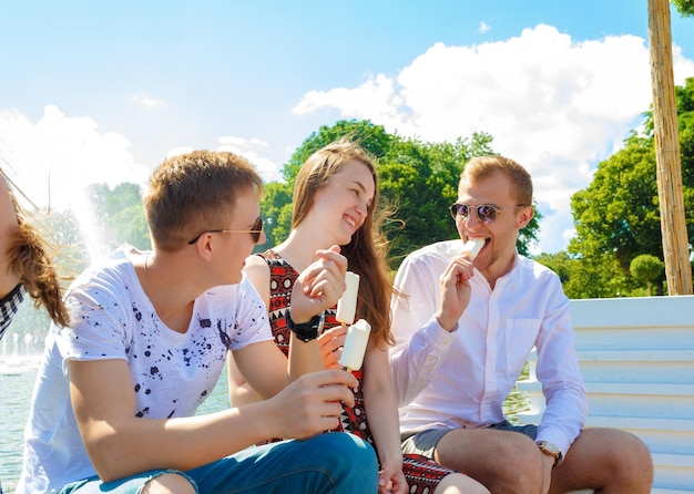 우정, 여가, 과자, 여름, 그리고 사람들의 개념 - 야외에서 아이스크림을 들고 웃고 있는 친구들. 화창한 여름날 공원에 있는 학생들은 벤치에 앉아 아이스크림을 먹고 있다