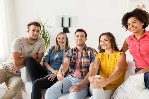 friendship, and people concept - group of happy friends taking picture with smartphone and selfie stick at home