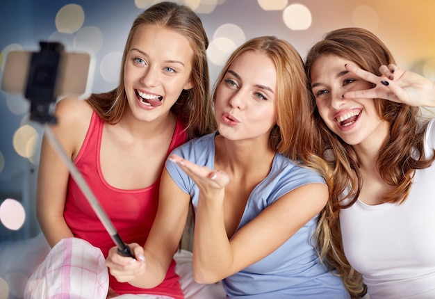 Foto amicizia gente festa pigiama e concetto di tecnologia amici felici o ragazze adolescenti con smartphone e monopod che si fanno selfie a casa