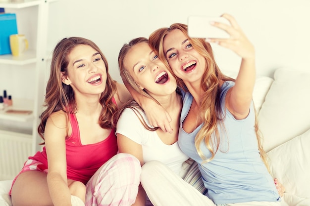 friendship, people, pajama party and technology concept - happy friends or teenage girls with smartphone taking selfie at home