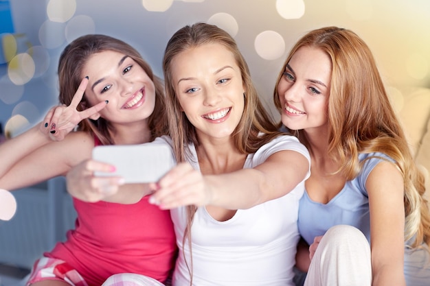 Foto amicizia persone festa di pigiama e concetto di tecnologia amici felici o ragazze adolescenti con smartphone che si fanno selfie a casa