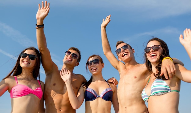 дружба, море, праздники, жест и концепция людей - группа улыбающихся друзей в купальниках и солнцезащитных очках, машущих руками на пляже
