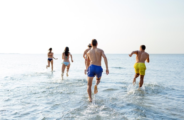 friendship, sea, summer vacation, holidays and people concept - group of smiling friends in swimwear running on beach from back