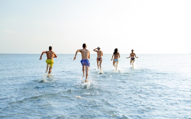 friendship, sea, summer vacation, holidays and people concept - group of smiling friends in swimwear running on beach from back