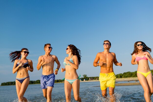 friendship, sea, summer vacation, holidays and people concept - group of smiling friends wearing swimwear and sunglasses running on beach