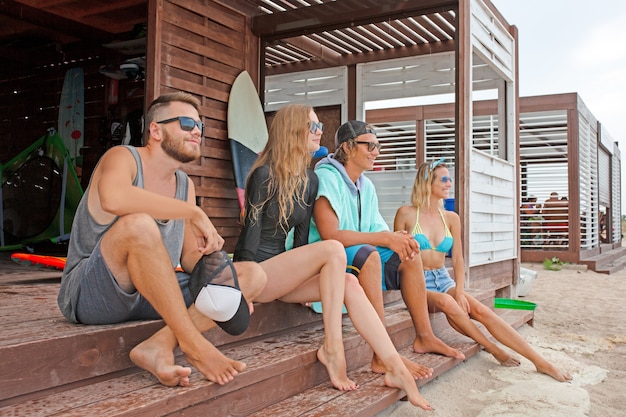 Friendship, sea, summer vacation, water sport and people concept - group of friends wearing swimwear sitting with surfboards on beach