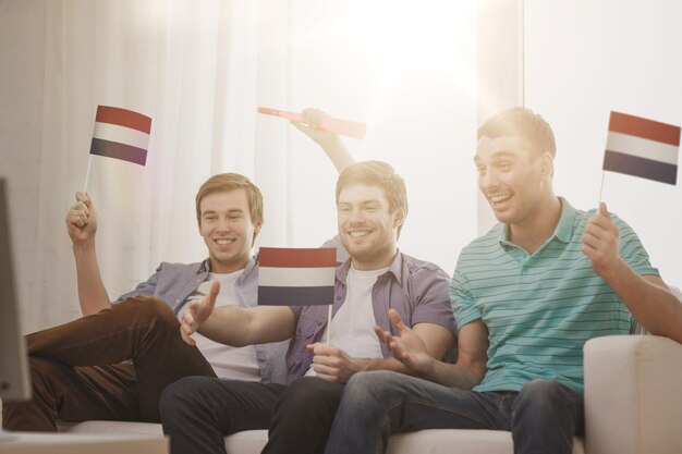 friendship, sports and entertainment concept - happy male friends with flags and vuvuzela supporting football team at home