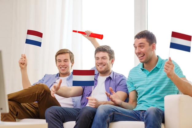 友情、スポーツ、エンターテインメントの概念-自宅でサッカーチームをサポートする旗とブブゼラを持つ幸せな男性の友人