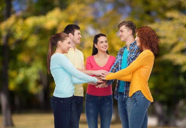 friendship, summer vacation, teamwork, gesture and people concept - group of smiling teenagers putting hand on top of each other over green park background