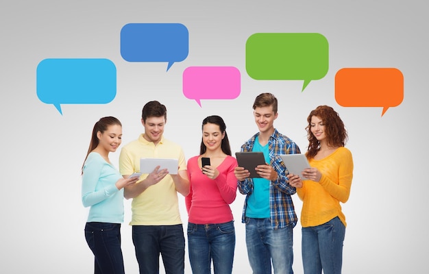 Photo friendship, technology, communication and people concept - group of smiling teenagers with smartphones and tablet pc computers over messenger text bubbles and gray background