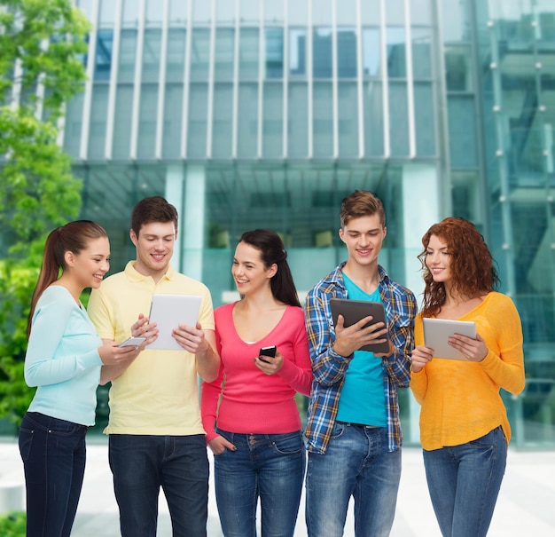 friendship, technology, education, business and people concept - group of smiling teenagers with smartphones and tablet pc computers over campus background