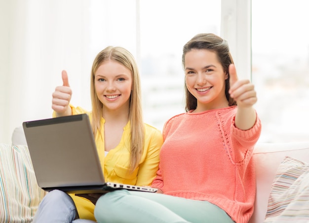 friendship, technology and internet concept - two smiling teenage girls with laptop computer at home showing thumbs up