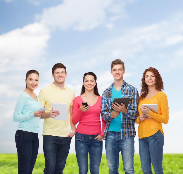 дружба, технологии, лето и концепция людей - группа улыбающихся подростков со смартфонами и планшетными компьютерами на фоне голубого неба и травы