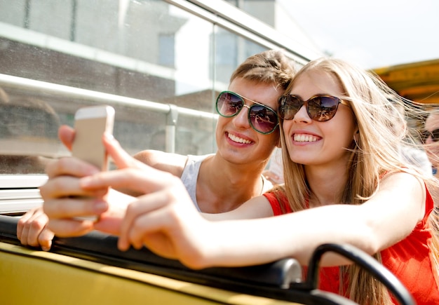 Photo friendship, tourism, summer vacation, technology and people concept - smiling couple with smartphone traveling by tour bus and making selfie