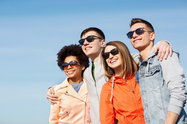 концепция дружбы, туризма, путешествий и людей - группа счастливых друзей-подростков в солнцезащитных очках, обнимающихся на открытом воздухе