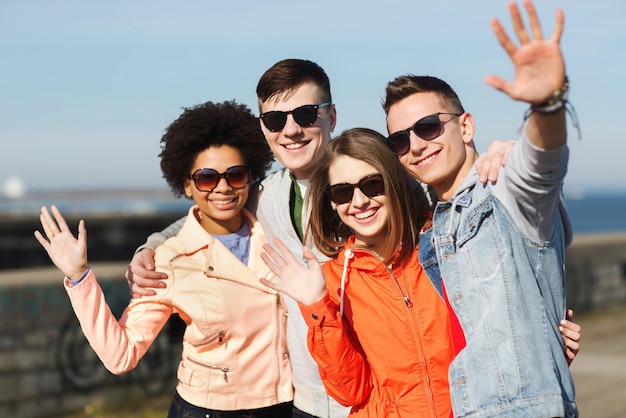 Concetto di amicizia, turismo, viaggi e persone - gruppo di amici adolescenti felici in occhiali da sole che abbracciano e agitano le mani all'aperto