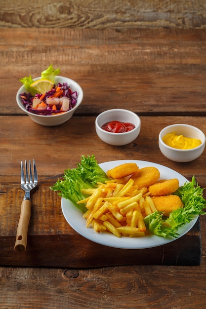 Frietjes met kipnuggets naast kaassaus en ketchup in een juskom op houten planken