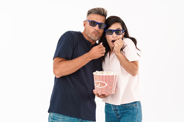 겁에 질린 매력적인 커플은 캐주얼 복장을 하고 흰 벽 위에 고립되어 서서 팝콘과 3d 안경으로 영화를 보고 있다