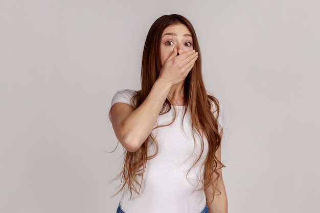Frightened dark haired woman covering mouth intimidated scared to talk, looking surprised terrified, keeping terrible secret, wearing white T-shirt. Indoor studio shot isolated on gray background.