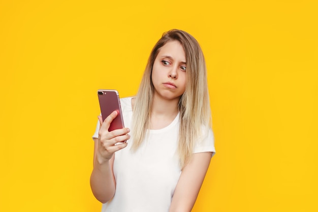 A frightened young caucasian blonde woman in a white tshirt with a mobile phone in her hand looks suspiciously at the incoming call from an unknown number on a bright color yellow wall