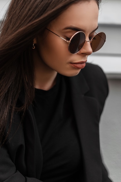 Fris portret van schattige mooie jonge vrouw in ronde modieuze zonnebril in zwarte stijlvolle blazer met sexy lippen buiten in de stad. aantrekkelijk meisje mannequin in casual elegante zakelijke kleding.