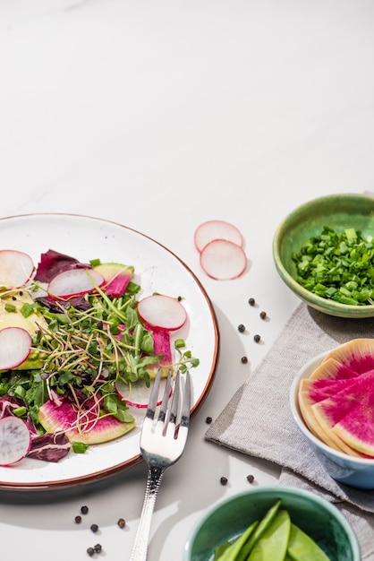 Frisse radijssalade met greens en avocado op plaat op witte ondergrond met ingrediënten in kommen en