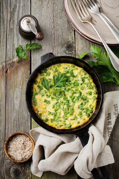 Foto fritatta con patate, piselli ed erbe aromatiche in una padella di ferro su una vecchia superficie di legno. messa a fuoco selettiva. stile rustico vista dall'alto.