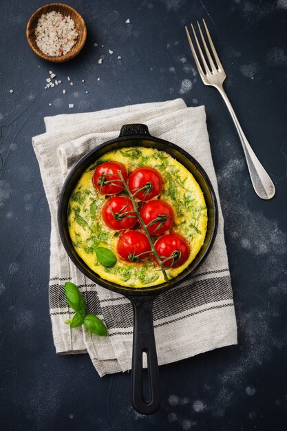 Frittata with arugula, potatoes and cherry tomatoes in iron pan on old stone table. Top view.