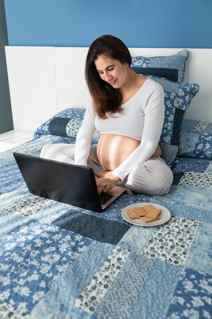 写真 自宅の快適なベッドに座ってネットブックを ⁇ 覧している笑顔の妊婦の上から