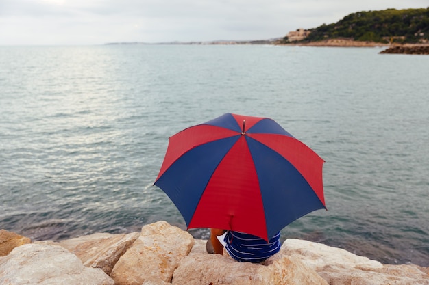 From behind anonymous man sitting on the rocks with an umbrella by the sea while it rains