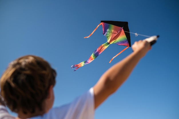 From below back view of unrecognizable kid with multicolored rainbow kite flying against cloudless blue sky in sunny summer day