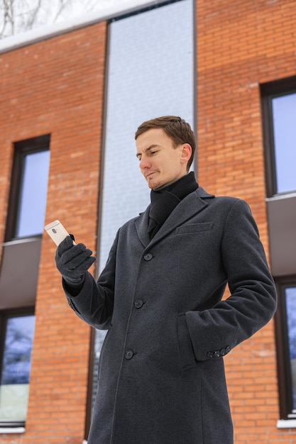 Снизу бизнесмен в зимнем пальто с помощью мобильного телефона против офисного здания на городской улице