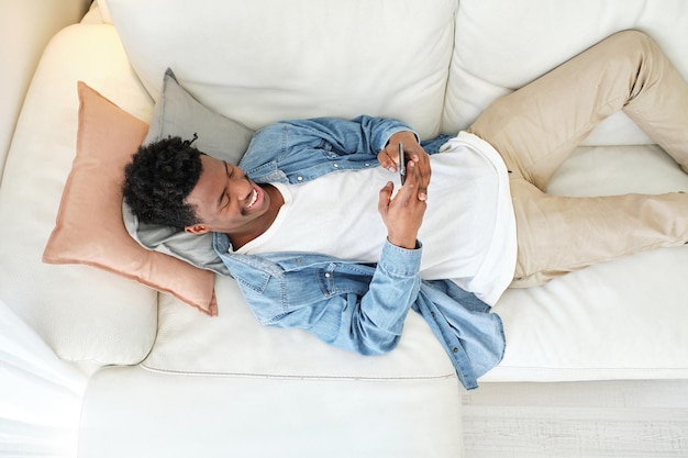 from above cheerful african american male in denim jacket relaxing on cozy couch and browsing mobile phone in light living room